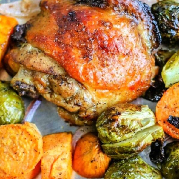 Sheet pan chicken thighs with charred skin, nestled among baked sweet potatoes, Brussels sprouts, and red onion on a baking sheet.