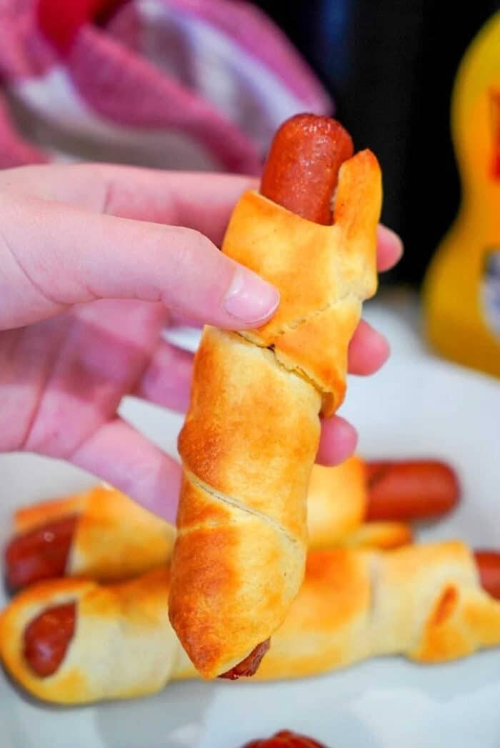 Best air fryer pigs in a blanket are my kids favorite snacks! Easy recipe to make with crescent rolls and hot dogs that turns out crunchy to perfection every time. A hand holding an air fryer pigs in a blanket, with more delicious crescent roll-wrapped hot dogs on a plate in the background.