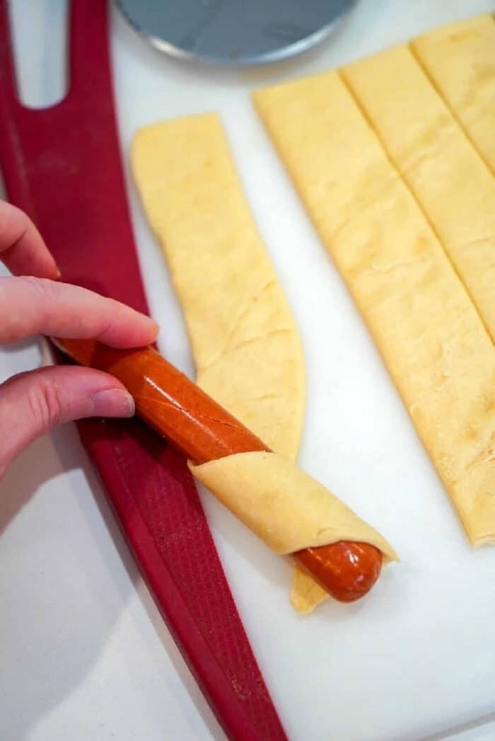 Easy air fryer pigs in a blanket are my kids favorite snacks! Easy recipe to make with crescent rolls and hot dogs that turns out crunchy to perfection every time. A hand wraps a sausage with a strip of dough on a cutting board, preparing delicious air fryer pigs in a blanket.