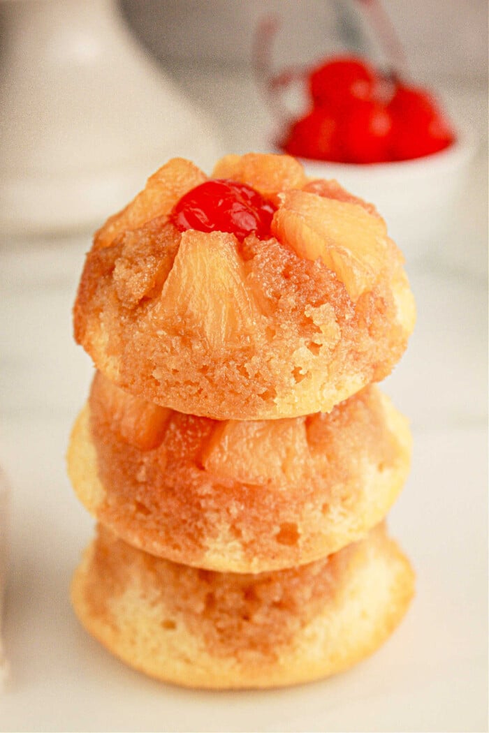 Three mini pineapple upside-down cupcakes are stacked on top of each other. Each cupcake features caramelized pineapple slices and a maraschino cherry on top. A small bowl of cherries is blurred in the background.