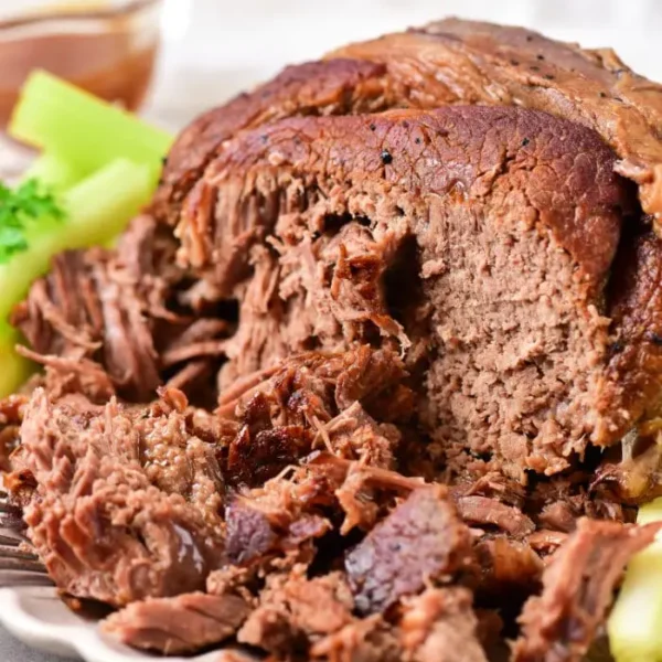 Close-up of a cooked beef roast shoulder on a white plate, surrounded by celery pieces. The roast is tender and slightly shredded, with a visible moist texture. A small bowl with sauce is in the background, perfectly complementing this delicious dish.