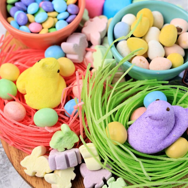 A wooden Candy Charcuterie Board displays colorful candies, including chocolate eggs, pastel marshmallow chicks, bunnies, and other shapes. The candies are arranged in nests of green and pink edible grass. Two bowls hold additional treats for delightful grazing.