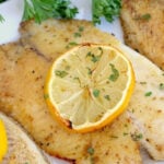 Seasoned frozen fish in air fryer fillet garnished with parsley and a slice of lemon, served on a white plate.