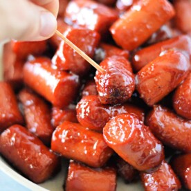 A hand holding a toothpick picks up a glazed mini sausage from a bowl of hot dog burnt ends.