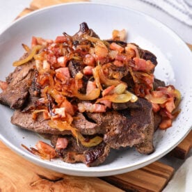 A white plate containing a dish of cooked liver topped with caramelized onions, diced tomatoes, and bits of bacon or ham is placed on a wooden board.