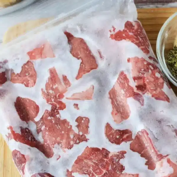 A clear plastic bag holds frozen pieces of red meat, perfect for those wondering how to cook frozen beef. On a wooden cutting board beside the bag is a small bowl with green herbs, resting on what seems to be a kitchen countertop.