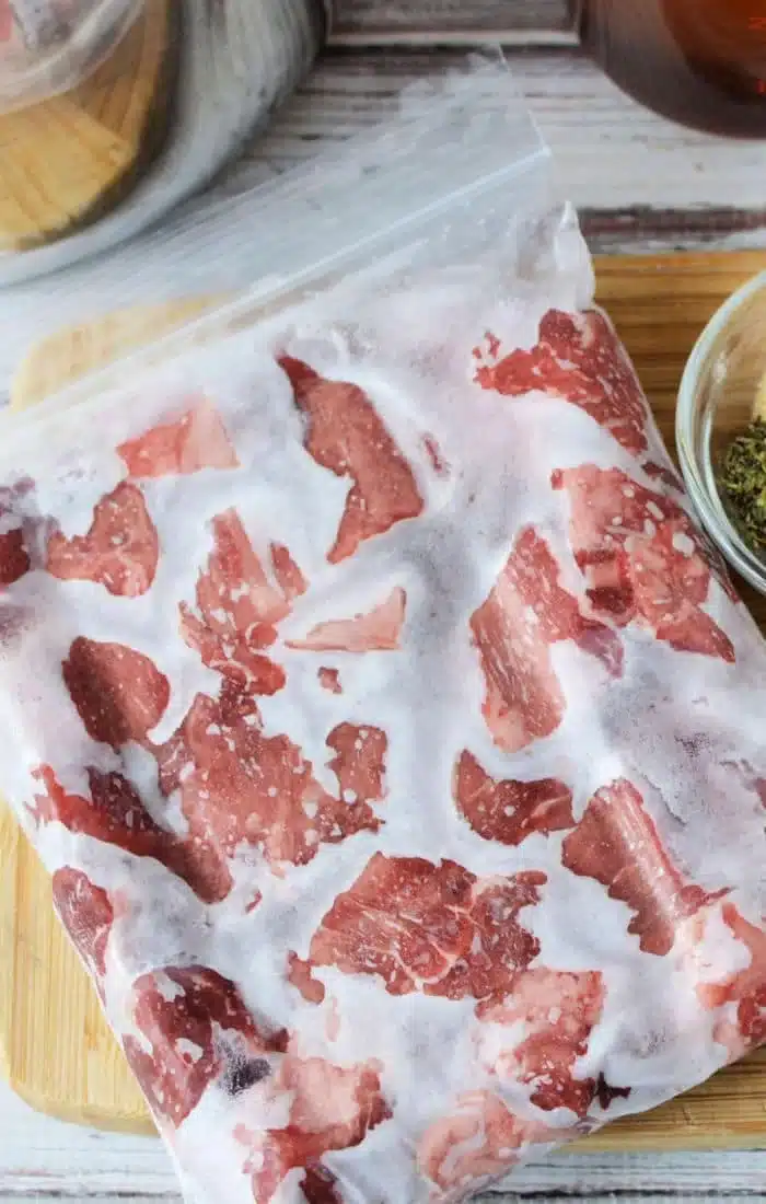 A clear plastic bag holds frozen pieces of red meat, perfect for those wondering how to cook frozen beef. On a wooden cutting board beside the bag is a small bowl with green herbs, resting on what seems to be a kitchen countertop.