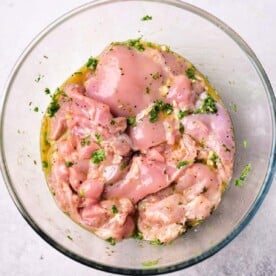 Raw, marinated chicken pieces, infused with a Mediterranean chicken marinade and herbs, rest in a clear glass bowl on a light surface.