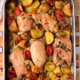 Mediterranean chicken thighs roasted with potatoes and cherry tomatoes in a metal pan on a wooden board.