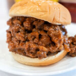 A sloppy joe sandwich with saucy ground meat filling on a sesame seed bun, perfectly paired with classic sloppy joe sides, served on a white plate.