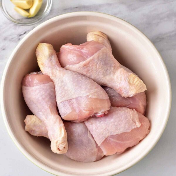 Raw chicken drumsticks in a beige bowl, accompanied by a small bowl of garlic cloves and a dish of mayonnaise on a marble surface, set the stage for crafting some of the best chicken leg recipes.