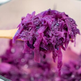 A wooden spoon lifts a serving of sautéed red cabbage above a pot brimming with more. The cabbage is soft and moist, showcasing a vibrant purple hue.