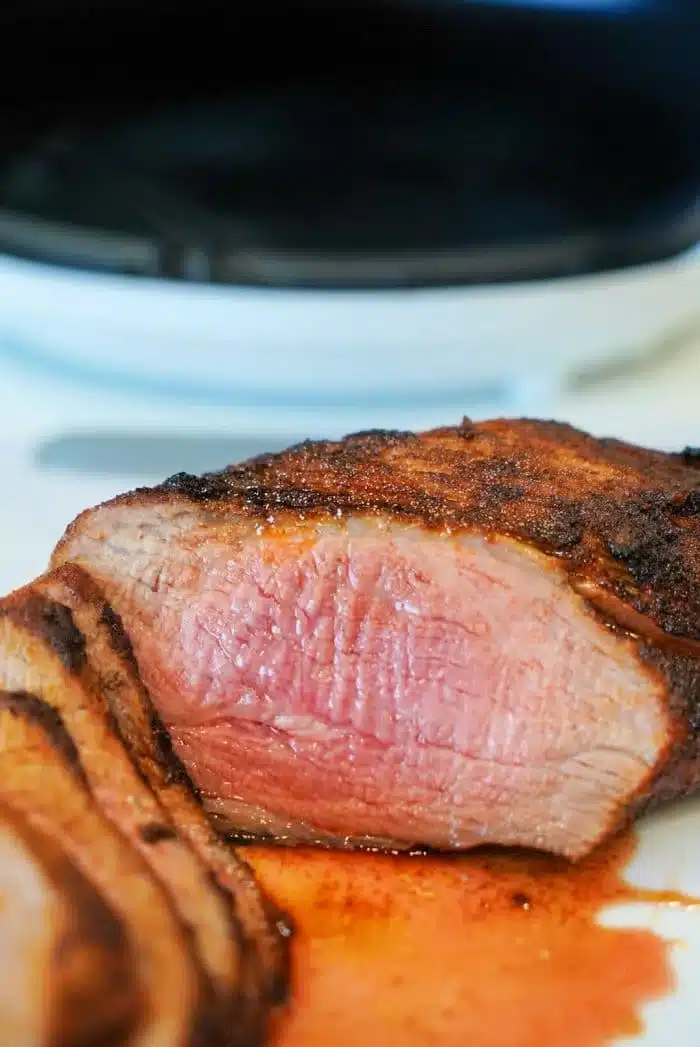 Close-up of sliced, air fryer-grilled tri tip with a medium-rare center. The steak is juicy, with a charred outer layer, resting on a white surface. A small amount of juice is pooled around the slices, highlighting the meat's tenderness.