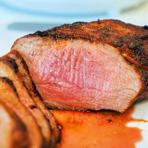 Close-up of a cooked air fryer tri-tip steak, sliced to reveal a pink, juicy interior. The steak boasts a seasoned, grilled crust with some juices pooled around it on the white plate.