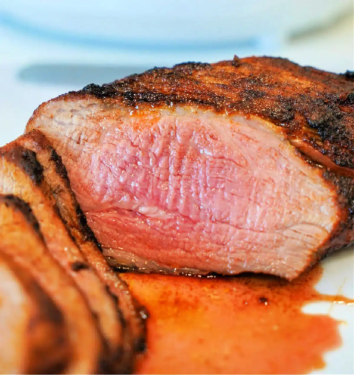 Close-up of a cooked air fryer tri-tip steak, sliced to reveal a pink, juicy interior. The steak boasts a seasoned, grilled crust with some juices pooled around it on the white plate.