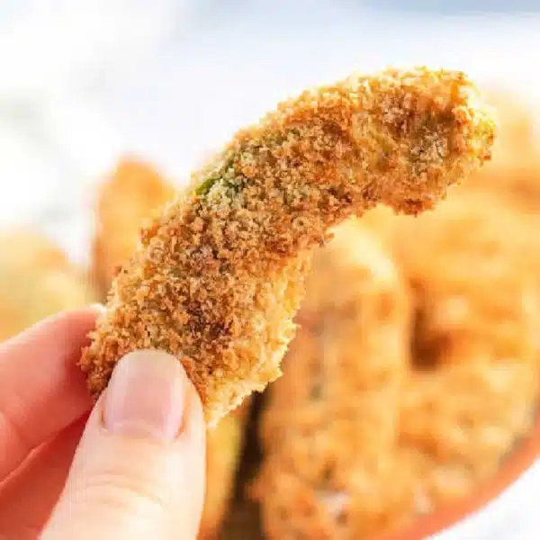 Close-up of a hand holding an air fryer avocado fry. The avocado is coated in a crispy, golden-brown crust, with more pieces in soft focus in the background.