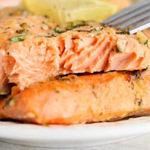 Close-up of a fork cutting into a perfectly cooked piece of salmon, garnished with herbs on a white plate. Follow steps on how to cook frozen salmon for this tender texture and flavor.