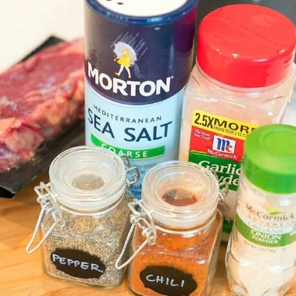 Assorted spices, including sea salt, garlic powder, pepper, chili, and minced onion—essentials for learning how to make fajita seasoning—with raw meat in the background.