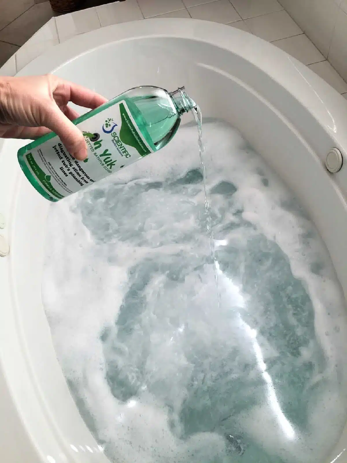 A hand pours liquid soap into a jetted tub filled with water and bubbles. The bottle, with its green label, complements the gleaming white oval-shaped bathtub as it effortlessly tackles the task of cleaning.