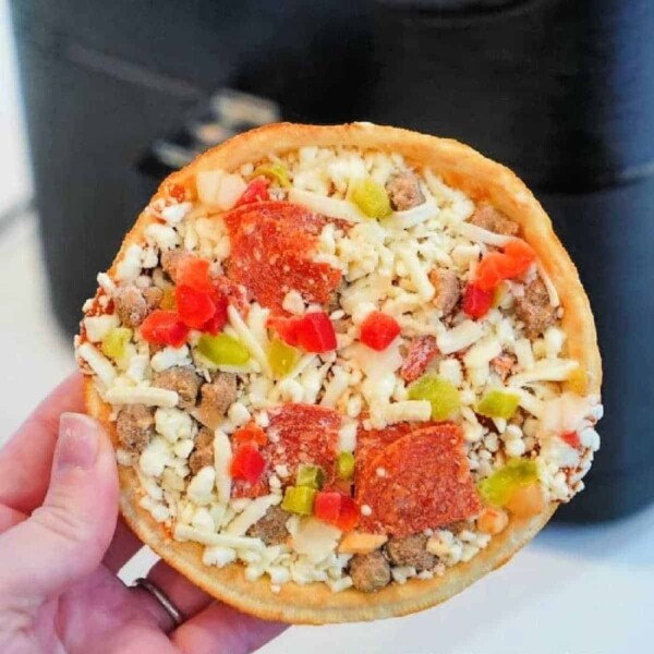 A hand holds a small, round frozen pizza, topped with diced green and red bell peppers, pepperoni slices, sausage crumbles, and shredded cheese. In the background is a black appliance—perfect for cooking frozen pizza in an air fryer.