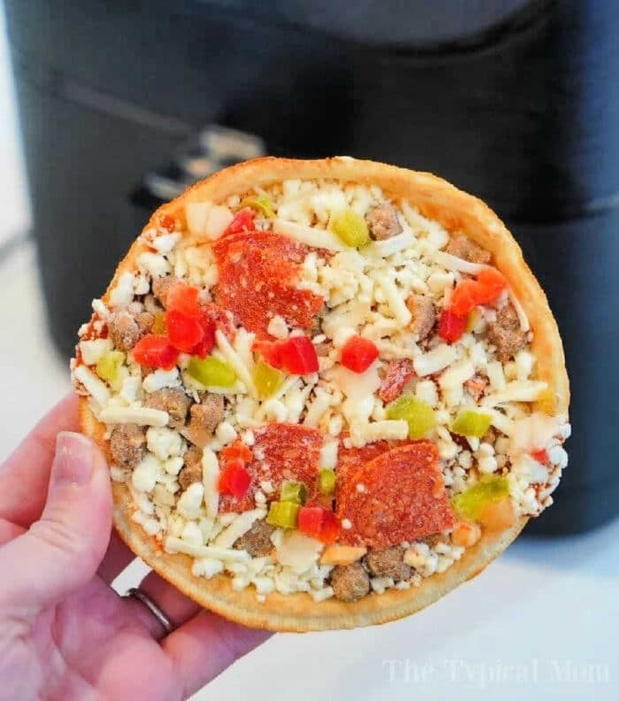 A hand holds a small, round frozen pizza, topped with diced green and red bell peppers, pepperoni slices, sausage crumbles, and shredded cheese. In the background is a black appliance—perfect for cooking frozen pizza in an air fryer.