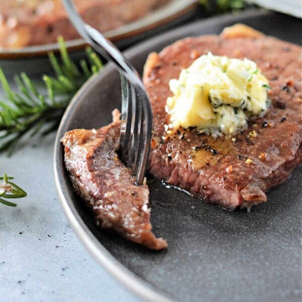 A perfectly cooked steak on a gray plate, topped with herb butter and sliced with a fork. Rosemary garnish complements the dish's presentation. Prepared to perfection using the Ninja Foodi Dual Zone Air Fryer for that ideal sear and tenderness.