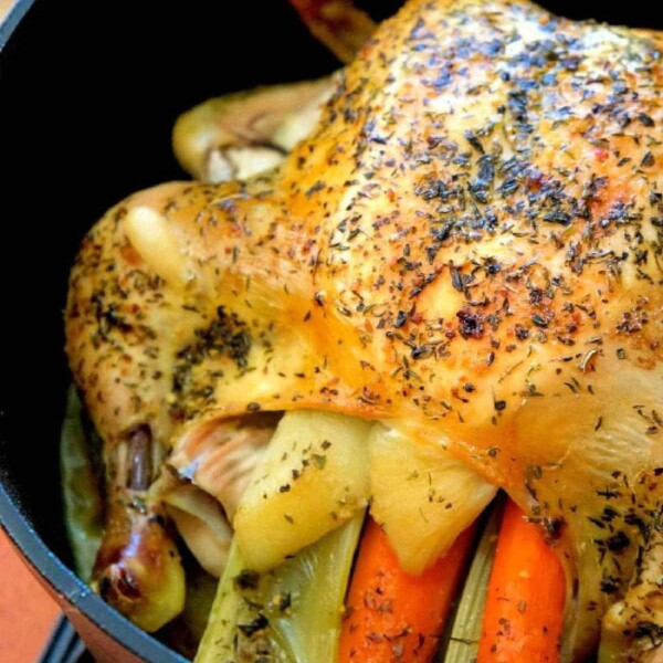 A seasoned whole chicken, richly infused with herbs, is nestled in a black Dutch oven. It's thoughtfully stuffed with carrots and celery, resting invitingly on a wooden surface.