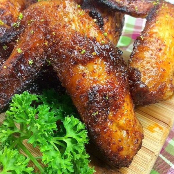 A close-up of crispy, golden brown Ninja Foodi chicken wings coated with seasoning on a wooden board. Fresh parsley is placed nearby, adding a pop of green. In the background, a red and white checkered cloth completes the rustic presentation.