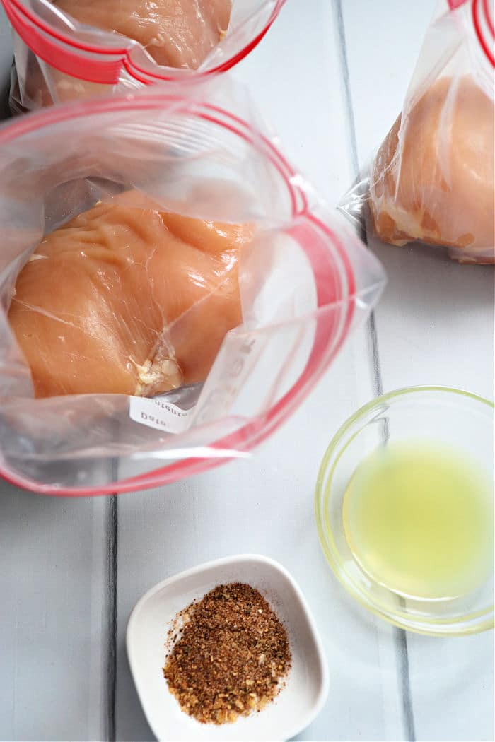 Raw chicken breasts in plastic bags, ready for how to sous vide chicken breast boneless, lie next to a small bowl of spice mix and a glass bowl of liquid on a white surface.