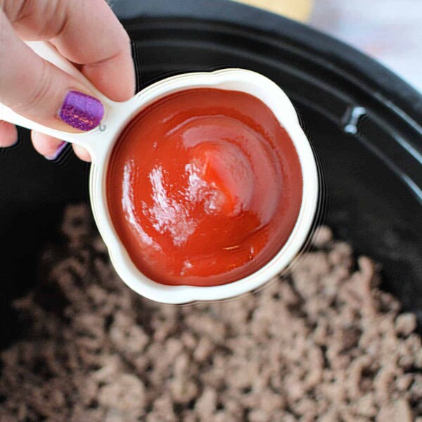 A hand holds a measuring cup of ketchup, poised above a pot of cooked ground meat—the beginning steps for crafting delicious slow cooker sloppy joes.