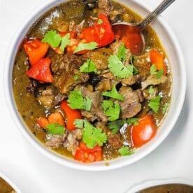 A bowl of hearty beef stew with chunks of tender beef, oxtail curry flavors, red tomatoes, and vibrant cilantro leaves, served with a spoon.