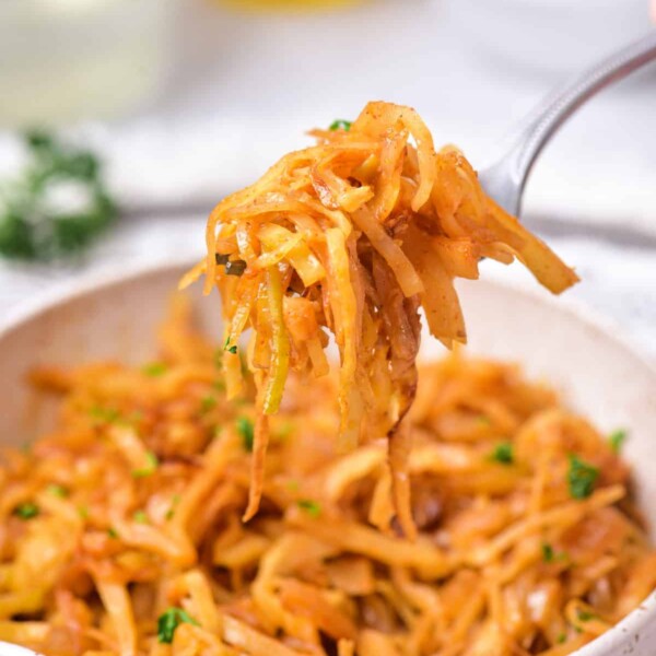 A fork holds a portion of sautéed shredded cabbage, perfectly embodying an Indian cabbage recipe, above a white bowl filled with more savory goodness. The dish is garnished with chopped herbs. In the blurred background, white bowls and clear bottles subtly enhance the scene.