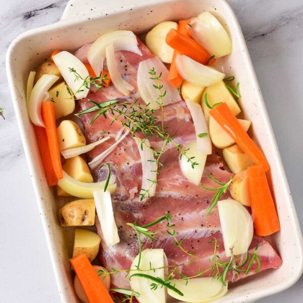 A baking dish holds raw chicken, sliced onions, carrots, and potatoes with fresh thyme sprigs on top. Nearby, a bowl of olive oil and a pepper mill rest on the marble surface, setting the stage for a comforting roast—much like contemplating how long to cook ribs in an oven at 350.