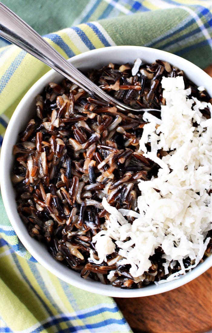 A delightful bowl of black rice pudding is topped with shredded coconut, resting invitingly on a plaid cloth alongside a spoon.