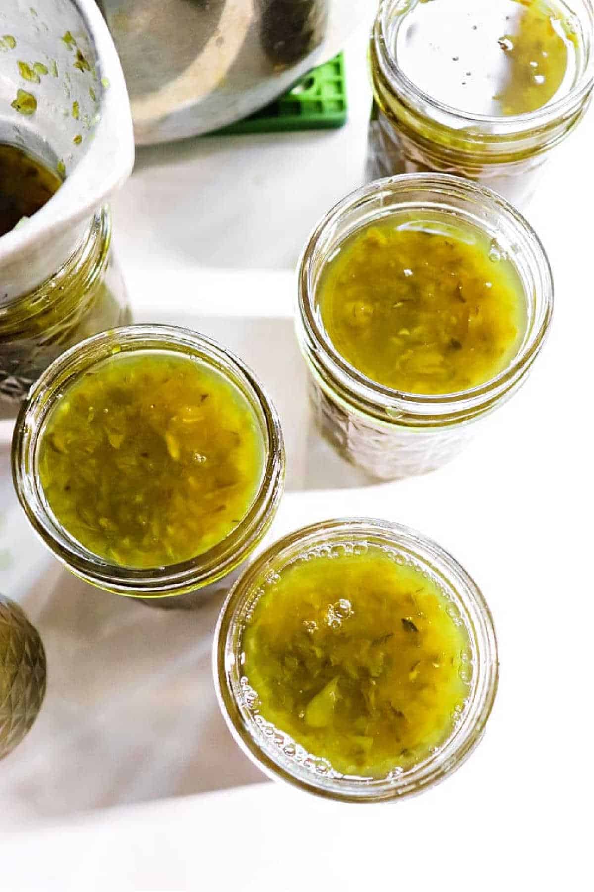 Four glass jars filled with homemade green relish and a hint of green Pepper Jelly sit on a white surface, ready for canning.