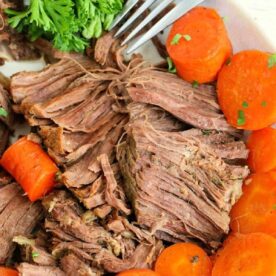 Close-up of a plate with shredded cooked beef, perfectly tender from being a frozen roast in the Instant Pot, surrounded by sliced, cooked carrots and a sprig of fresh parsley. A fork is placed beside the beef, ready to serve.