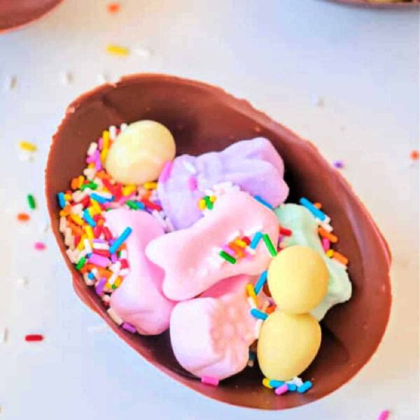 A chocolate surprise egg half filled with colorful marshmallows, yellow candies, and rainbow sprinkles rests on a white surface adorned with scattered sprinkles.