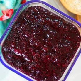 A square container filled with rich huckleberry jam sits next to a piece of bread and a colorful floral cloth.