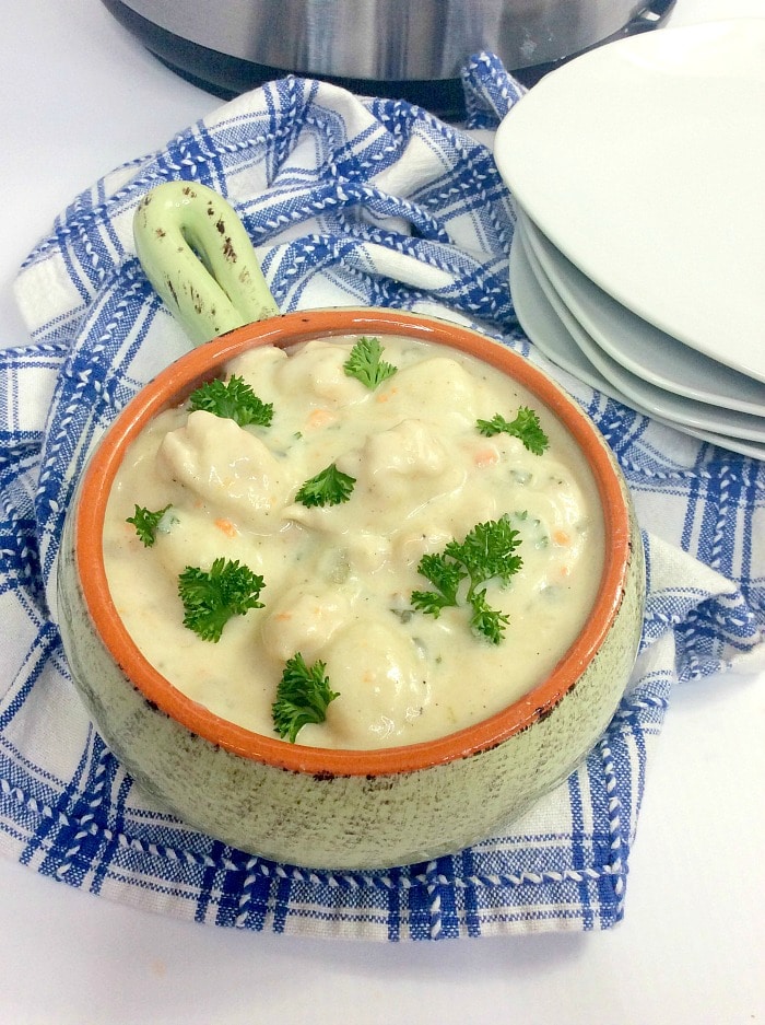 A bowl of creamy instant pot gnocchi soup with chunks of vegetables and parsley garnish, placed on a blue and white checkered cloth beside stacked white plates.