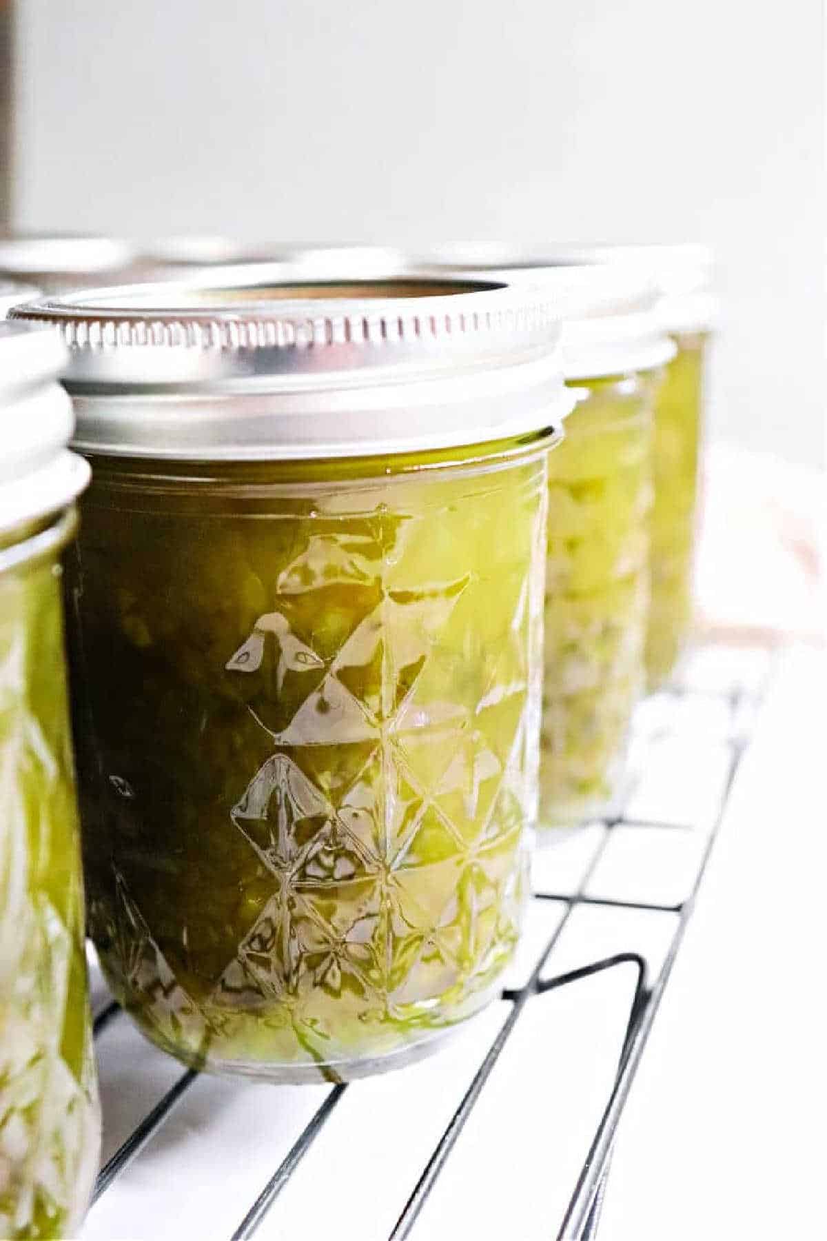 Close-up of jars filled with green Pepper Jelly and relish, sealed with metal lids, cooling on a wire rack.