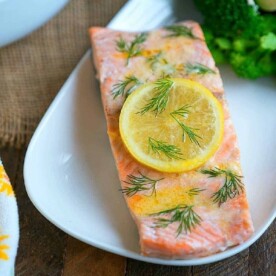 Salmon fillet garnished with a lemon slice and dill on a white plate, showcasing one of our delightful salmon dinner recipes, accompanied by broccoli in the background.