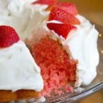 The delightful 2 ingredient strawberry cake features white frosting and is adorned with sliced strawberries on top. Presented elegantly on a glass platter, it tempts with a piece already missing.