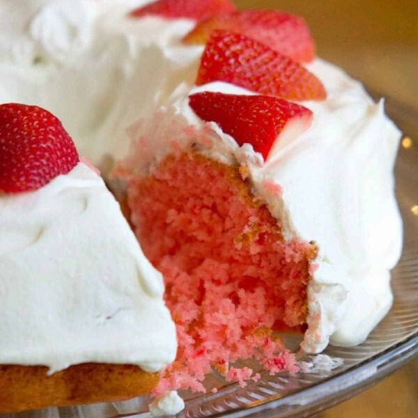 The delightful 2 ingredient strawberry cake features white frosting and is adorned with sliced strawberries on top. Presented elegantly on a glass platter, it tempts with a piece already missing.