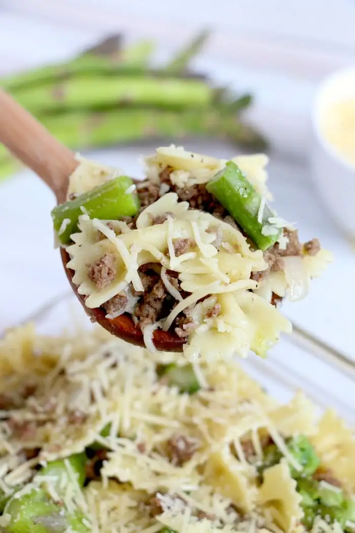 A wooden spoon holds a serving of bow-tie pasta mixed with ground meat, crisp bacon pieces, green asparagus, and topped with grated cheese over a dish of the same pasta. Blurred in the background are whole asparagus stalks.