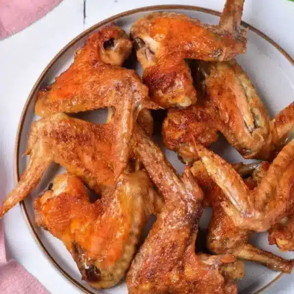 A plate of baked whole chicken wings is displayed on a white surface with a pink cloth and a few green herbs scattered around. The wings are golden-brown and appear crispy.