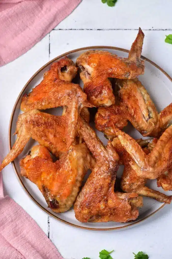 A plate of baked whole chicken wings is displayed on a white surface with a pink cloth and a few green herbs scattered around. The wings are golden-brown and appear crispy.