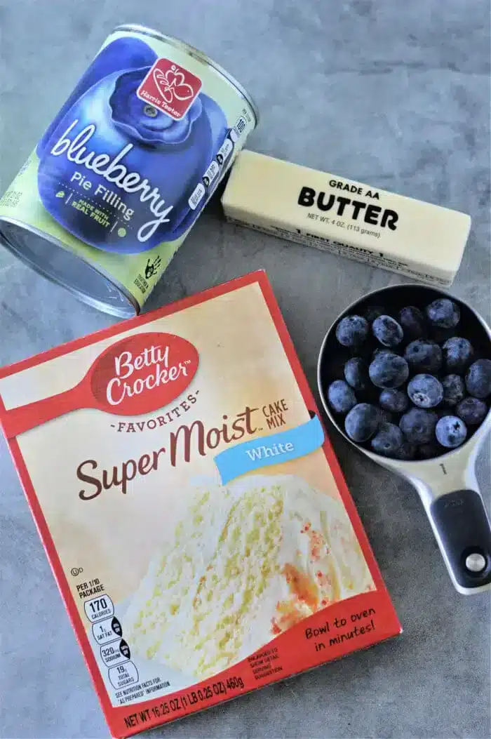 A can of blueberry pie filling, a stick of Grade AA butter, and a box of Betty Crocker Super Moist white cake mix are perfectly arranged on a gray surface, ready to create a delicious blueberry cobbler with cake mix. A measuring cup filled with fresh blueberries completes the scene.