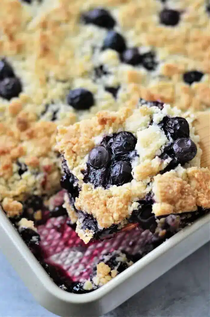 A close-up of a blueberry cobbler with cake mix in a baking pan. A portion is being lifted by a spatula, revealing layers of crumbly topping and juicy blueberries. The dessert is golden brown with visible purple-blue filling, merging the best of both worlds in a single bite.