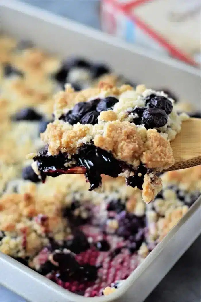 A close-up of a wooden spoon holding a serving of blueberry cobbler with cake mix, showcasing a golden, crumbly topping in a baking dish. Beneath the topping, juicy blueberries peek through enticingly.