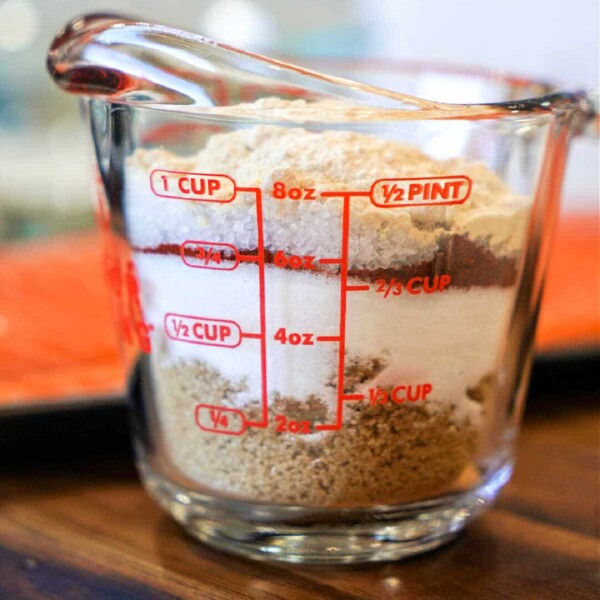 A glass measuring cup filled with layered ingredients, perfect for crafting a dry rub for chicken wings, including white sugar, brown sugar, and cocoa powder. Red measurement lines indicate ounces, cups, and pints on its clear surface. It rests elegantly on a wooden countertop.